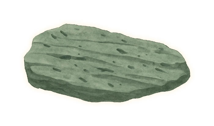 the same pale grey-green layered rock as the Isua sediment beside it on this page — a cool sage grey-green overall, with no warm buff or brown in it — scattered with dull mid-grey graphite flecks — short angular slivers and smears lying along the layering, like pencil lead rubbed into stone, never round bubbles or holes